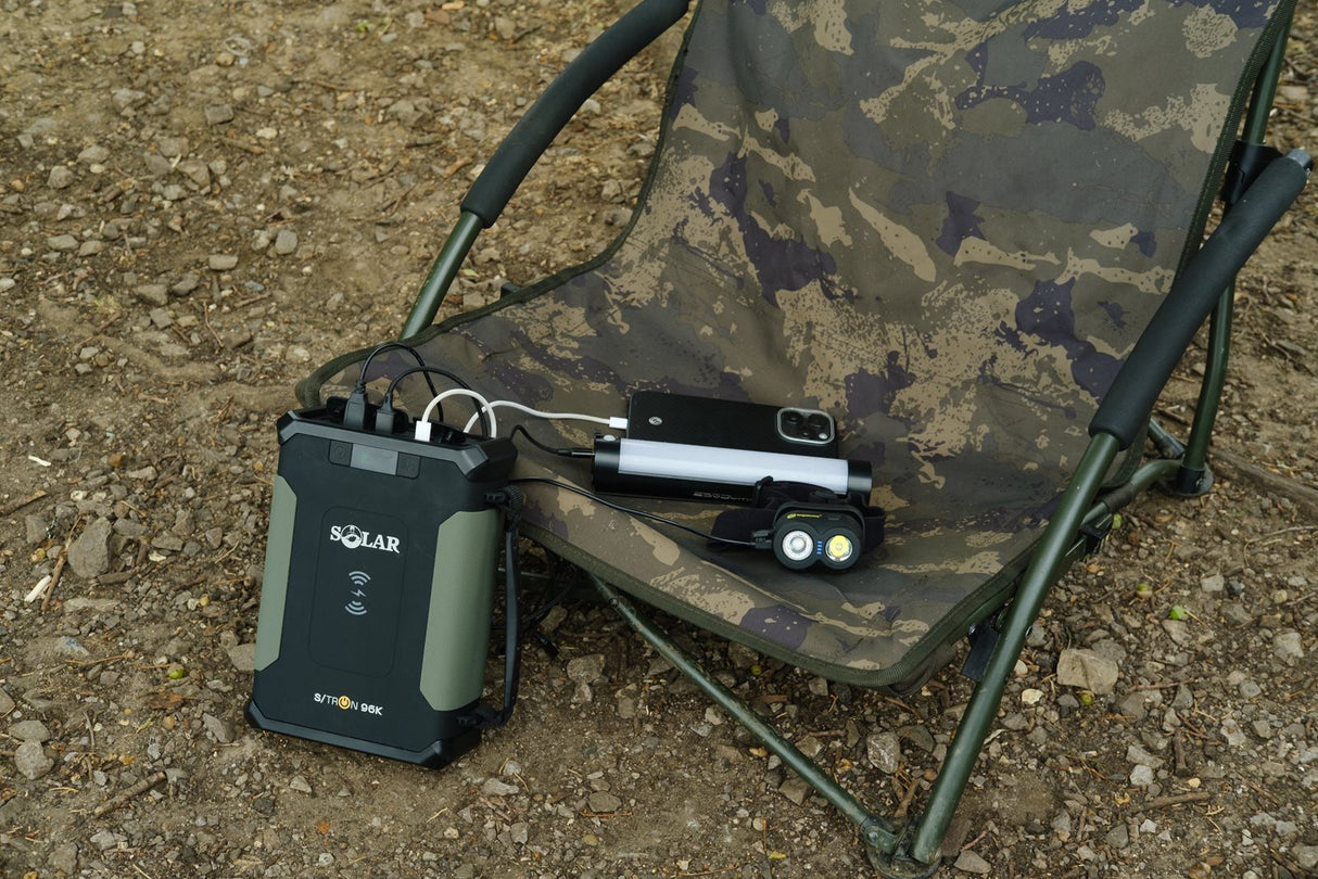 Solar S-Tron 96K powerpack charging a phone, a bivvy light and a headtorch beside a camo chair