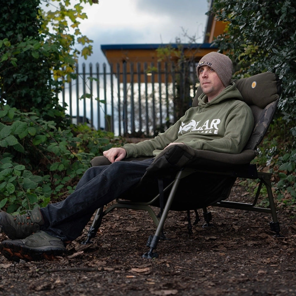 Lifestyle: an angler sitting back in the chair by a lake, showing the reclined position and the seat width in use. Same fleece headrest and camo back panel as the studio shot.