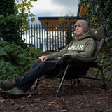 Lifestyle: an angler sitting back in the chair by a lake, showing the reclined position and the seat width in use. Same fleece headrest and camo back panel as the studio shot.