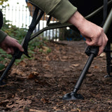 Close-up of a hand extending one of the spring-loaded telescopic legs onto its self-levelling swivel mud foot on leaf litter - the one-touch leg adjustment described in the copy.